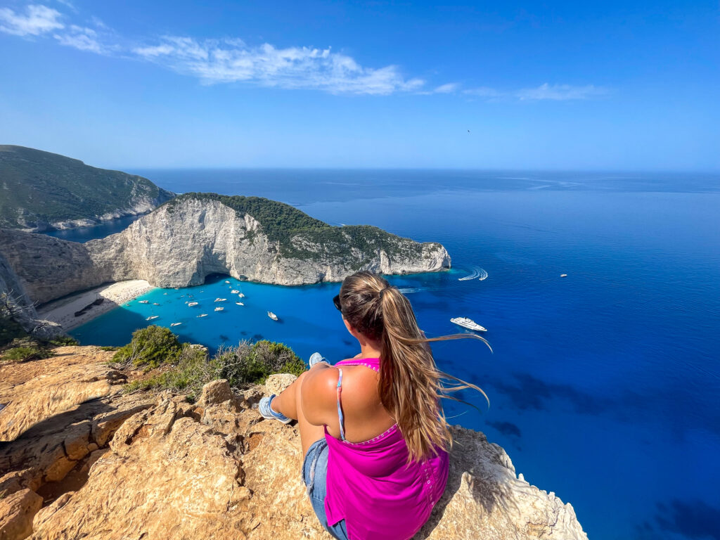 Hannah sitting on a cliff edge with her hair blowing in the wind as she looks down at shipwreck beach in Zakynthos