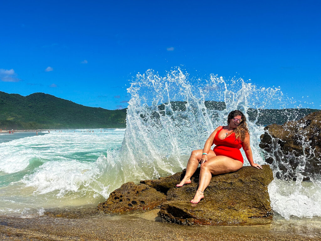 Hannah Logan in a red swimsuit sitting on a rock with a huge wave splashing behind her