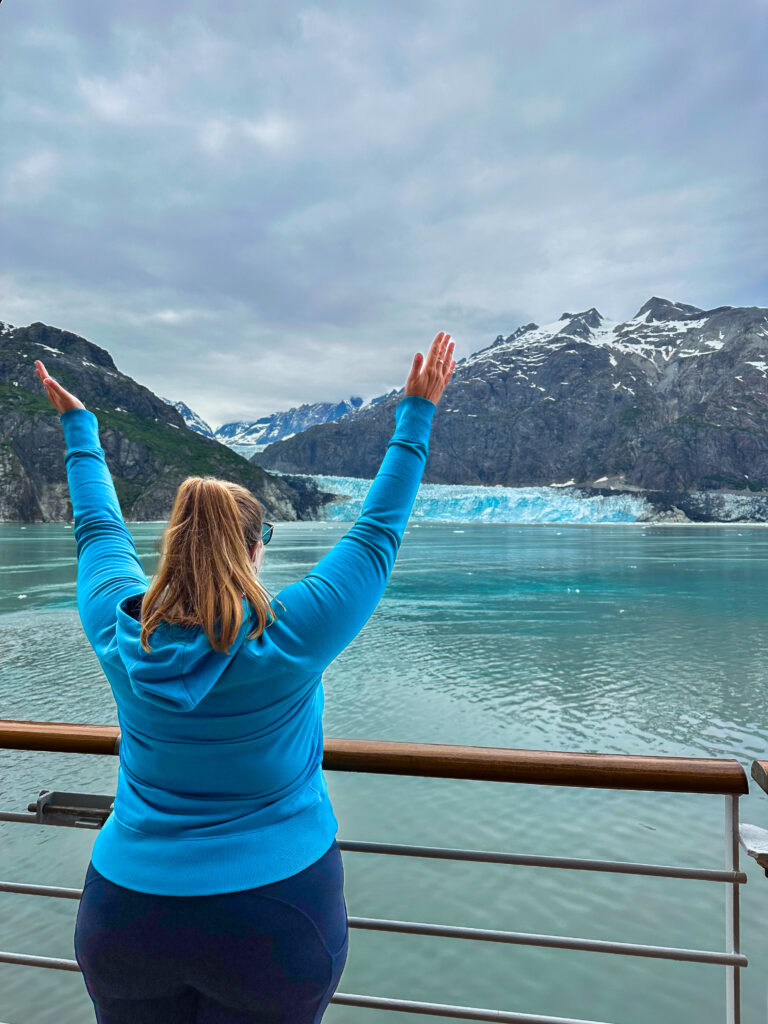 Hannah in a blue sweatshirt and her arms up in the air looking out at Margerie Glacier, Alaska