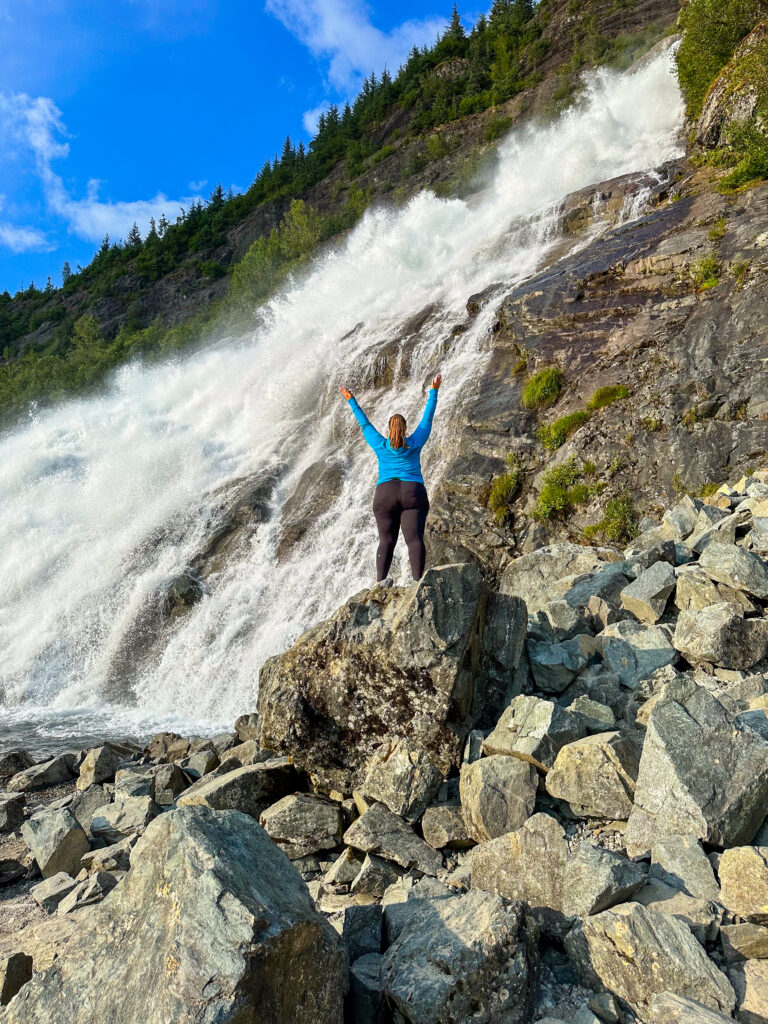 Hannah standing on a pile of rocks in front of a waterfall with her arms up in the air