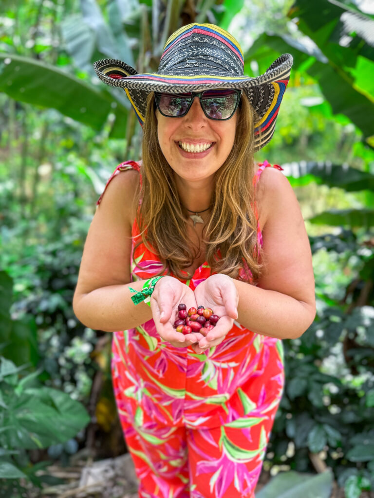 Hannah wearing a farming hat and bright pink jumpsuit holding a handful of coffee beans