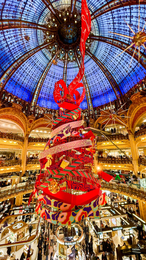 Huge Christmas tree in Paris shopping mall under beautiful glass dome ceiling. The tree is primarily red and gold with large Santas, elves, and other decorations.
