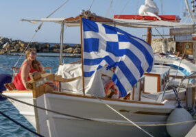Hannah in a red dress sitting on a boat with a Greek flag waving in the wind